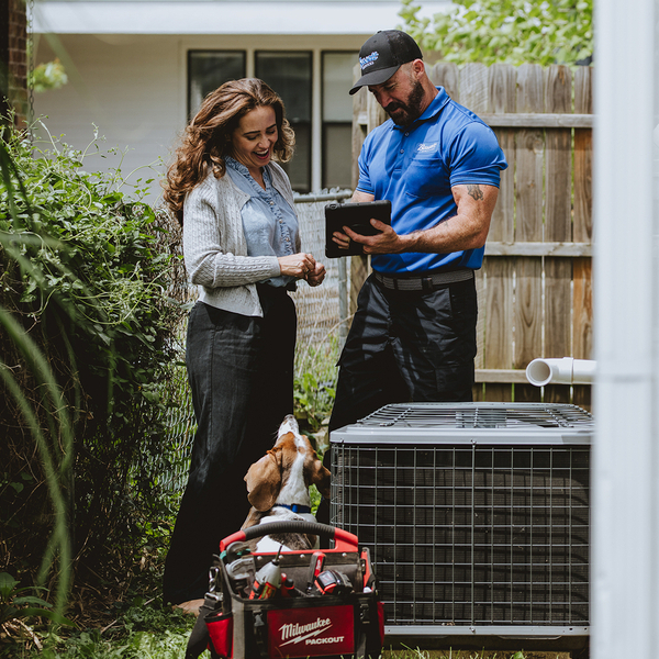Bassett Services technician showing a tablet to a homeowner beside her outdoor AC unit, with a basset hound nearby