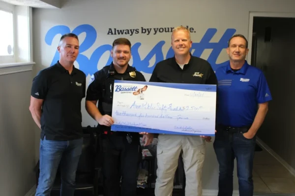 Bassett Services team members presenting a large donation check in front of the Bassett Always by your side logo wall